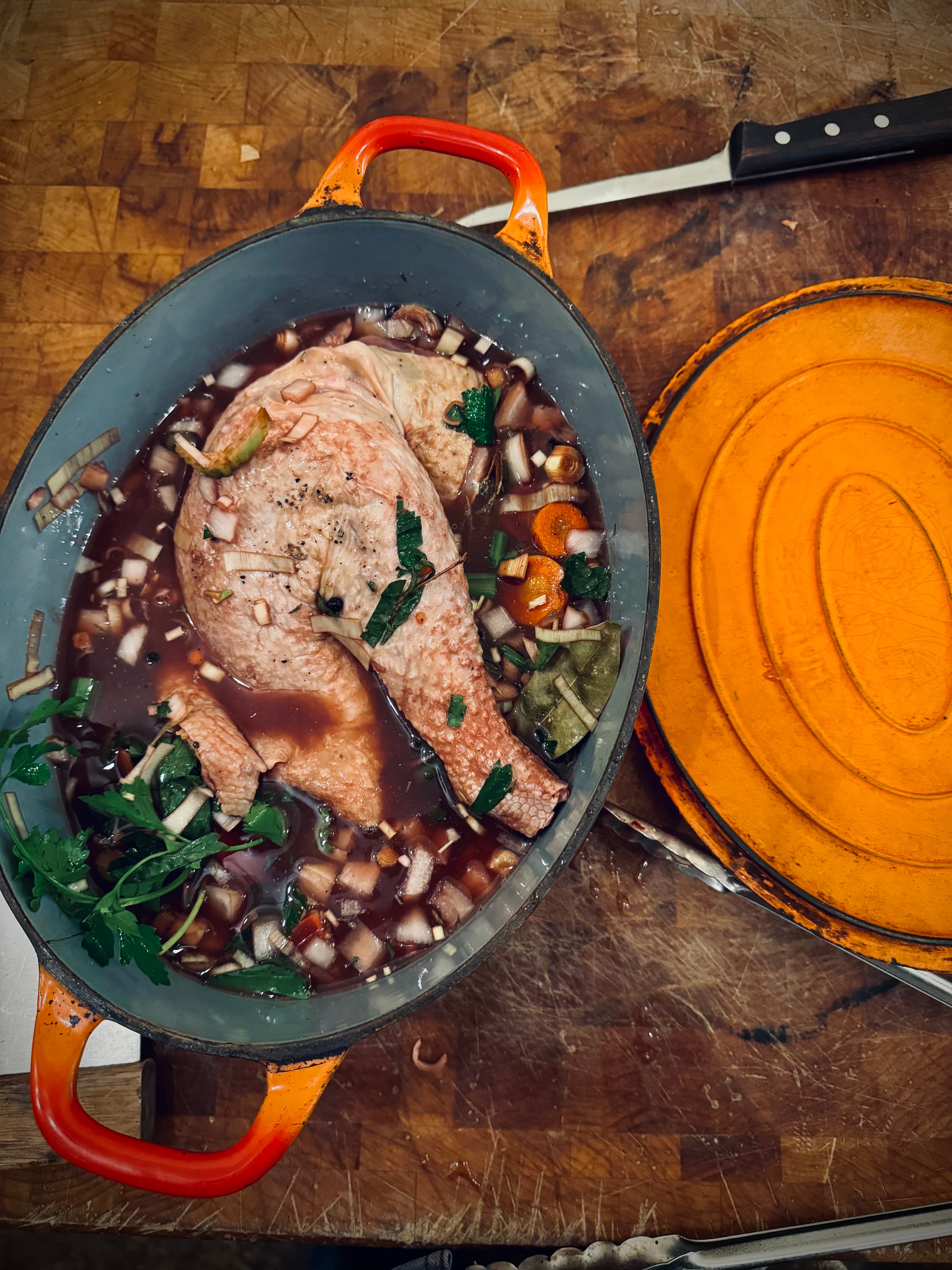 Rooster pieces marinating in farm wine with carrots, onions, and herbs in an orange Le Creuset casserole